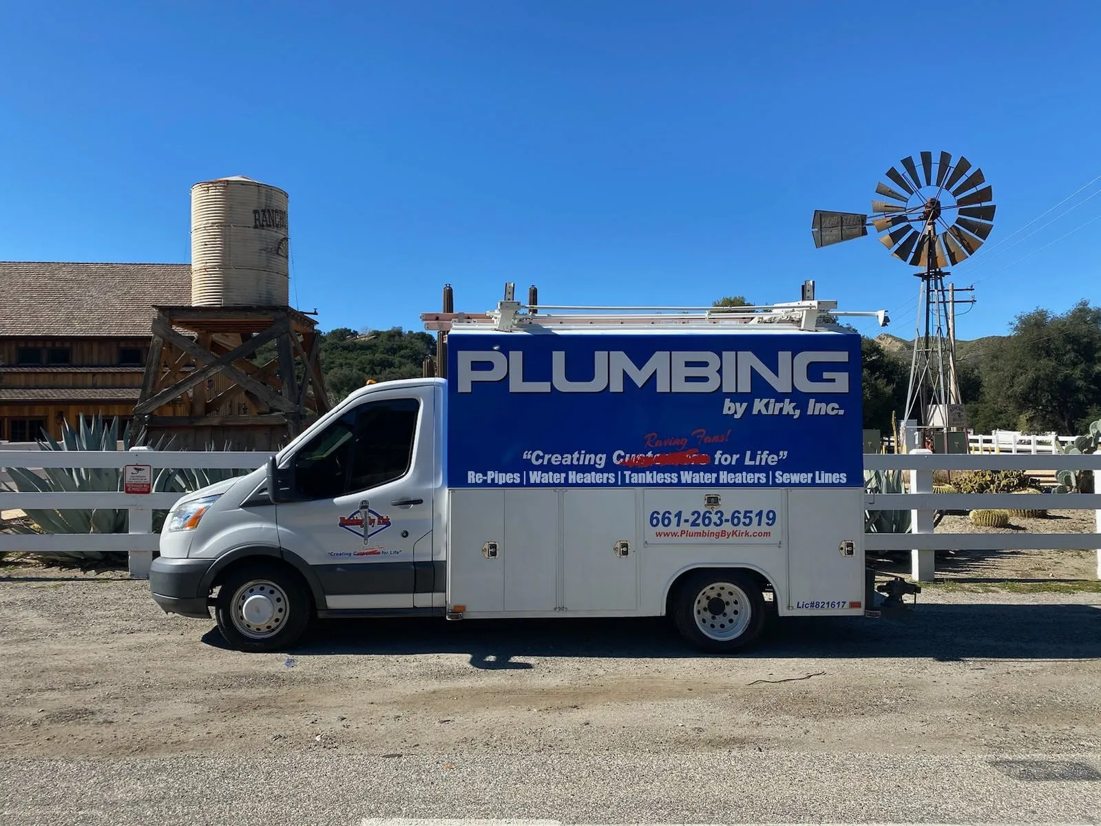 A Plumbing by Kirk service truck outside a ranch property with a windmill and a water tower